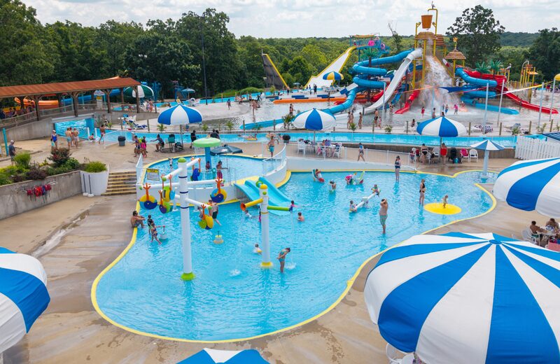 Shallow kiddie splash pad with colorful play structures, mini slides, and blue umbrellas