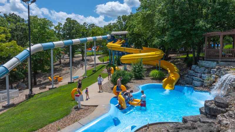 Yellow and blue twisting tube water slides ending in a bright blue splash pool with riders and a lifeguard